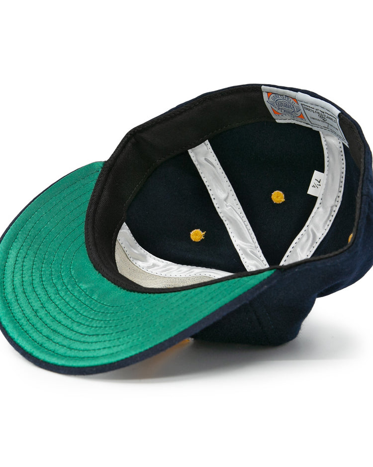 An authentic EFF Great Lakes Naval Station 1944 Vintage Ballcap, navy wool, is shown upside down to reveal its green underside brim, white lining, yellow rivets, visible labels, and size tag inside the crown.