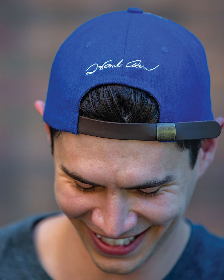 A smiling person wears the Ebbets Field Flannels Hank Aaron 1973 Signature Series Ballcap backwards, revealing white cursive writing on the back as they look down, with a blurred background behind them.