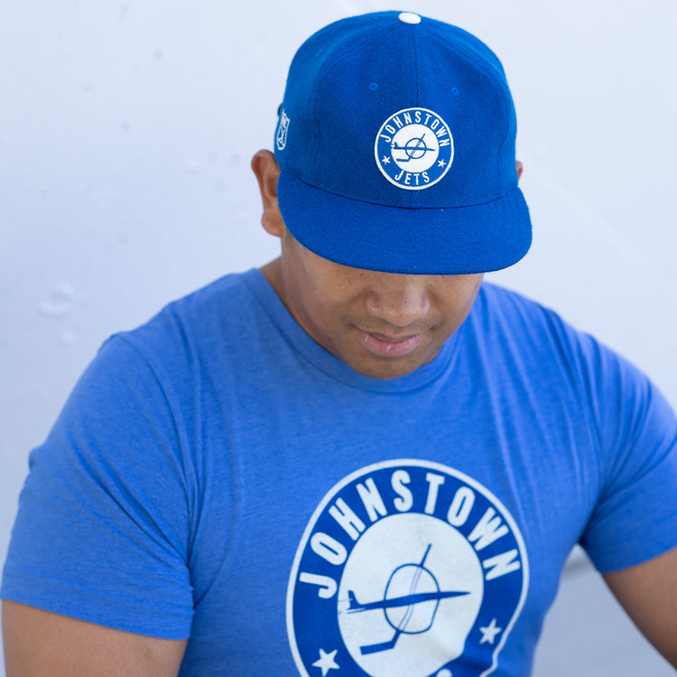 A man in an EFF Johnstown Jets 1970 Vintage Ballcap in blue, with an adjustable leather strap, and a matching blue circular-logo Johnstown T-shirt, looks down against a light background.