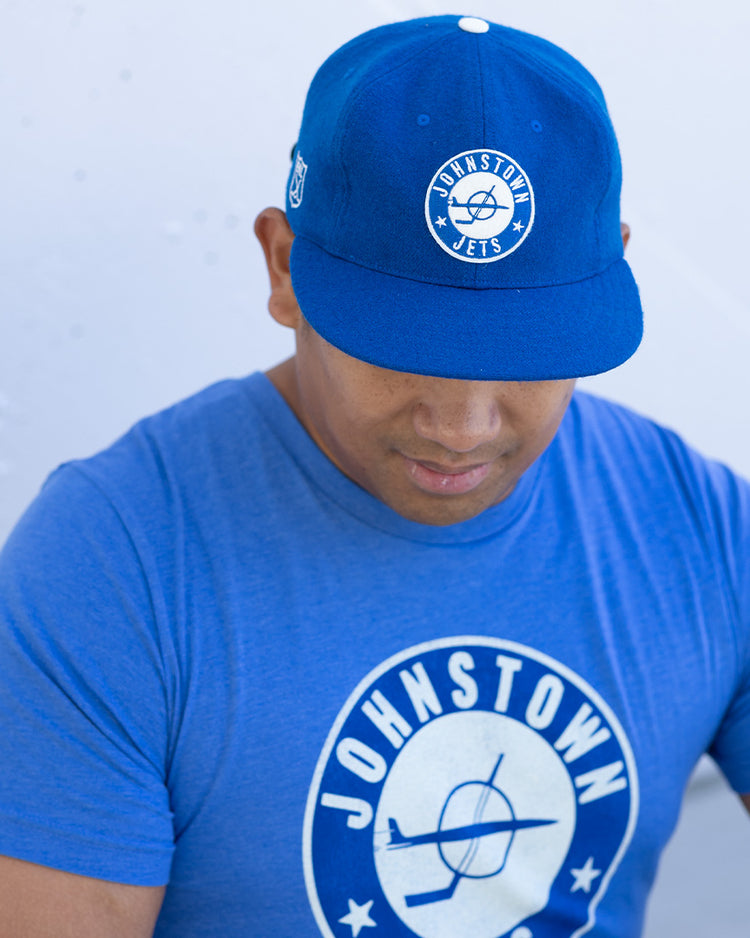 A man in an EFF Johnstown Jets 1970 Vintage Ballcap in blue, with an adjustable leather strap, and a matching blue circular-logo Johnstown T-shirt, looks down against a light background.