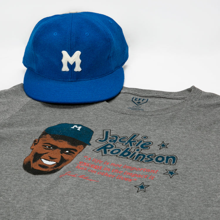 A blue baseball cap with a white M and an Ebbets Field Flannels Jackie Robinson Illustration T-Shirt—featuring his smiling portrait, name, and quote—are displayed on a white background.