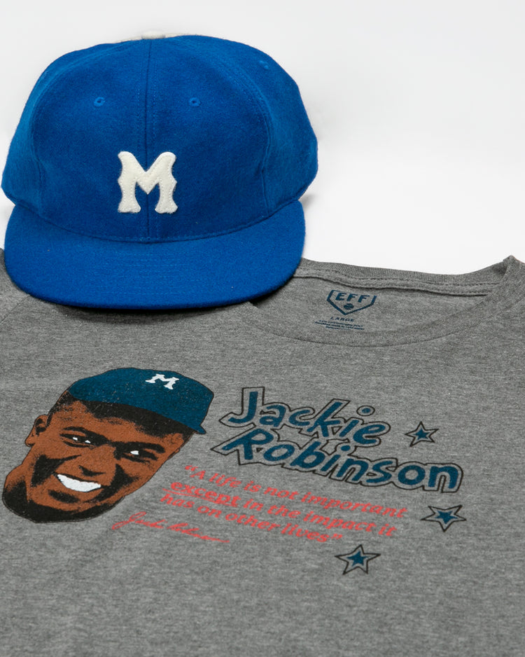 A blue baseball cap with a white M and an Ebbets Field Flannels Jackie Robinson Illustration T-Shirt—featuring his smiling portrait, name, and quote—are displayed on a white background.