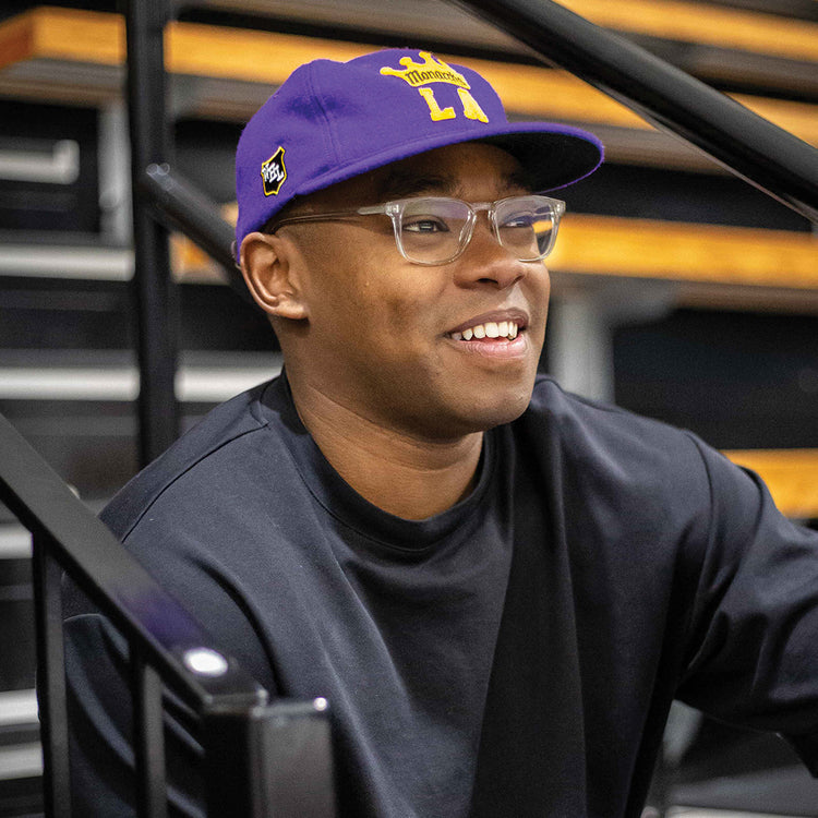 A smiling person wears the EFF Los Angeles Monarchs 1947 Vintage Ballcap, glasses, and a black shirt while sitting on bleachers with metal railings and wooden steps in the background.