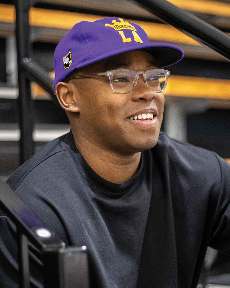 A smiling person wears the EFF Los Angeles Monarchs 1947 Vintage Ballcap, glasses, and a black shirt while sitting on bleachers with metal railings and wooden steps in the background.