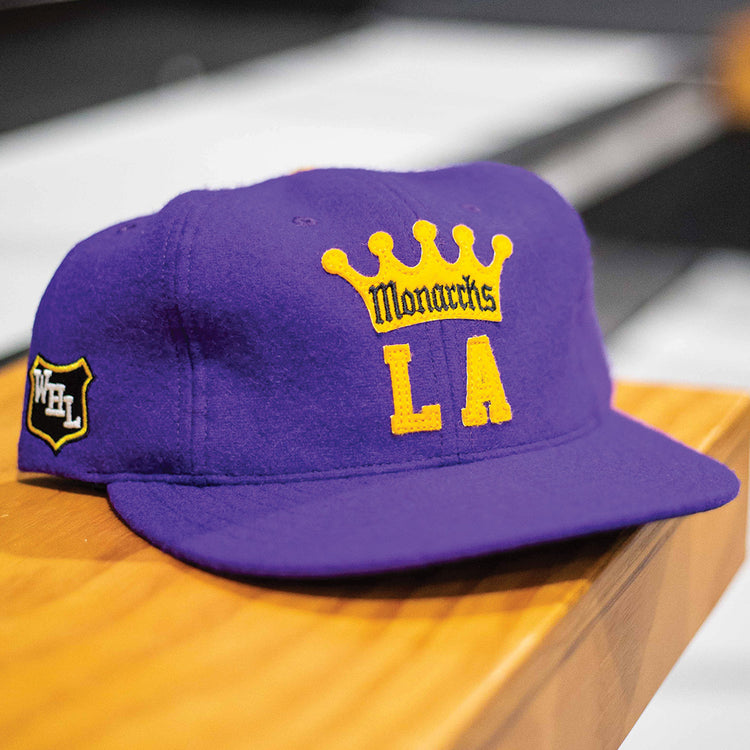 The EFF Los Angeles Monarchs 1947 Vintage Ballcap, featuring a purple base, yellow crown, Monarchs LA front embroidery, and a white-and-black Western Hockey League patch on the side, rests on a wooden surface.