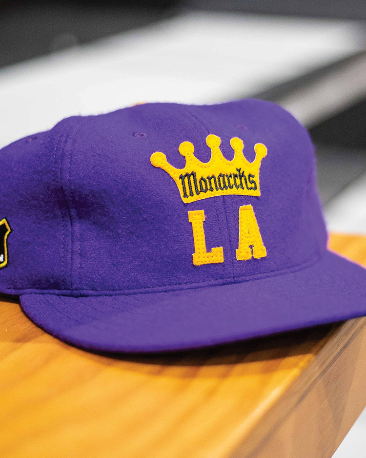 The EFF Los Angeles Monarchs 1947 Vintage Ballcap, featuring a purple base, yellow crown, Monarchs LA front embroidery, and a white-and-black Western Hockey League patch on the side, rests on a wooden surface.