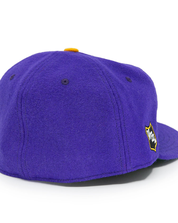 The EFF Los Angeles Monarchs 1947 Vintage Ballcap is a purple wool cap with a yellow top button, small ventilation holes, and a yellow and black shield logo on the side, shown from the back-left angle.