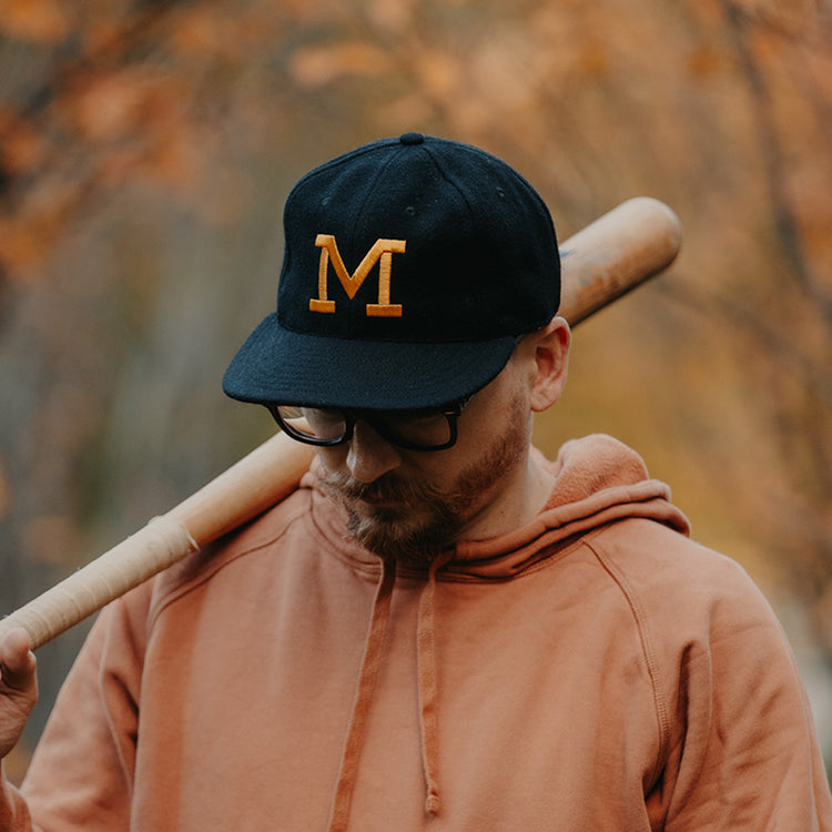 A man in an EFF University of Missouri 1958 Vintage Ballcap with a yellow M, glasses, and an orange hoodie rests a baseball bat on his shoulder, looking down amid a glowing autumn background.