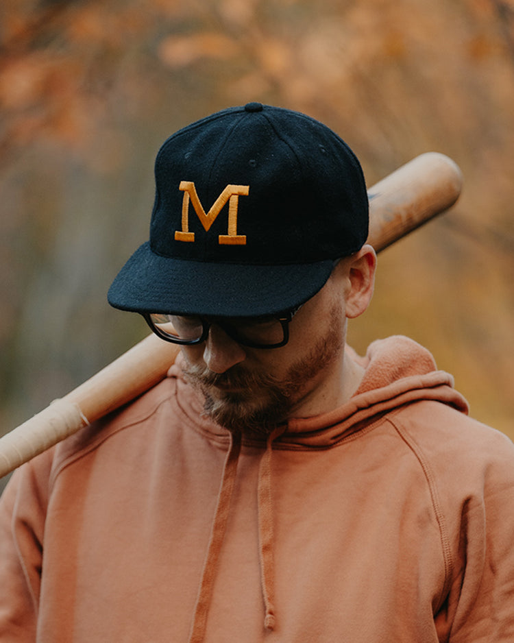 A man in an EFF University of Missouri 1958 Vintage Ballcap with a yellow M, glasses, and an orange hoodie rests a baseball bat on his shoulder, looking down amid a glowing autumn background.