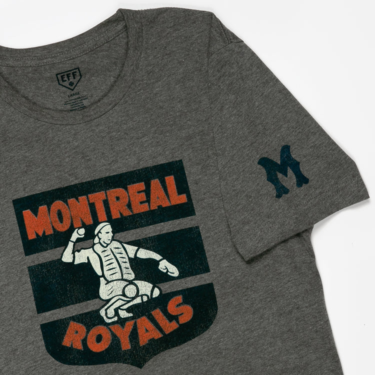 The EFF Montreal Royals 1946 T-Shirt is a gray tee with a graphic of a catcher in gear, Montreal Royals text in orange and cream, and a large blue M on the left sleeve—honoring Dodgers prospect Roberto Clemente.