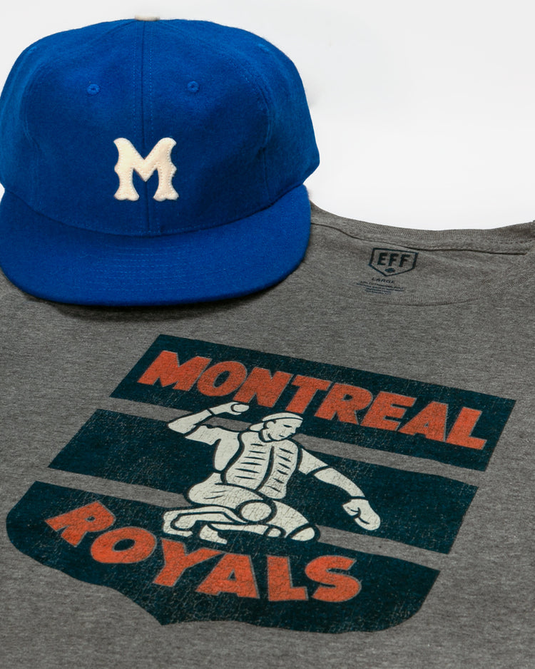 A blue baseball cap with a white M rests atop a gray EFF Montreal Royals 1946 T-Shirt, featuring a vintage-style catcher graphic and Dodger prospect roots.