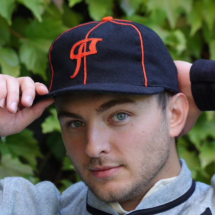 A young man with light eyes and stubble adjusts an EFF Montreal Black Panthers 1936 Vintage Ballcap, featuring orange stitching and logo, while standing before leafy green foliage.