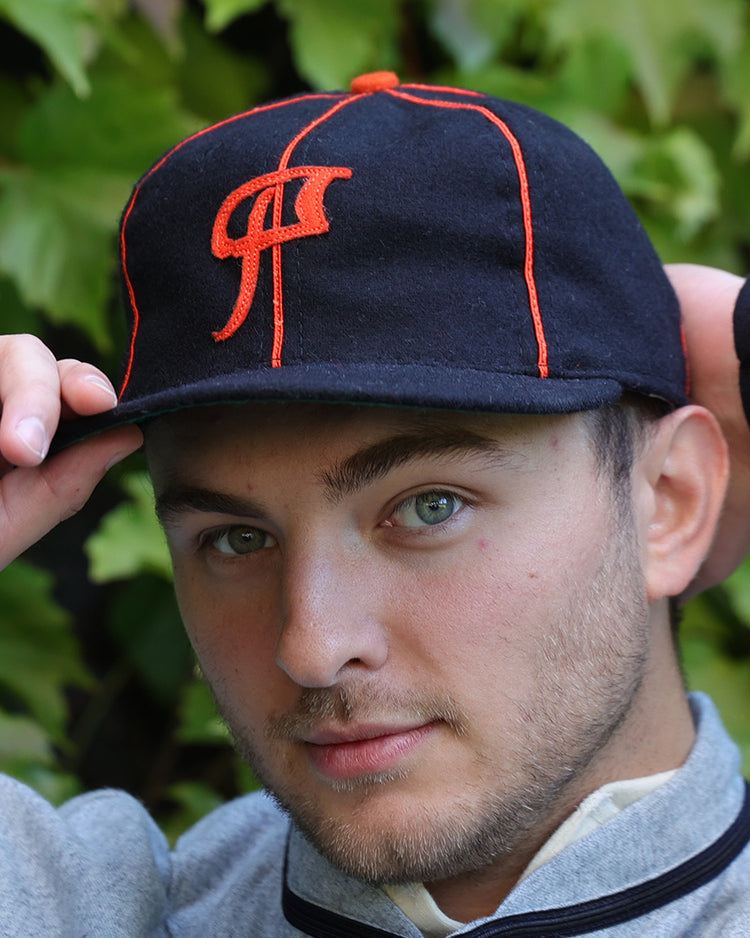 A young man with light eyes and stubble adjusts an EFF Montreal Black Panthers 1936 Vintage Ballcap, featuring orange stitching and logo, while standing before leafy green foliage.
