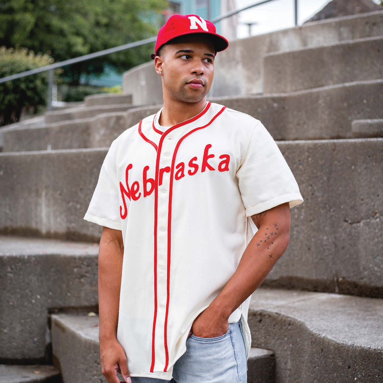 A man stands outdoors on concrete steps, looking to the side with one hand in his pocket, wearing an Ebbets Field Flannels University of Nebraska 1958 Vintage Ballcap and a white baseball jersey with Nebraska written on it.