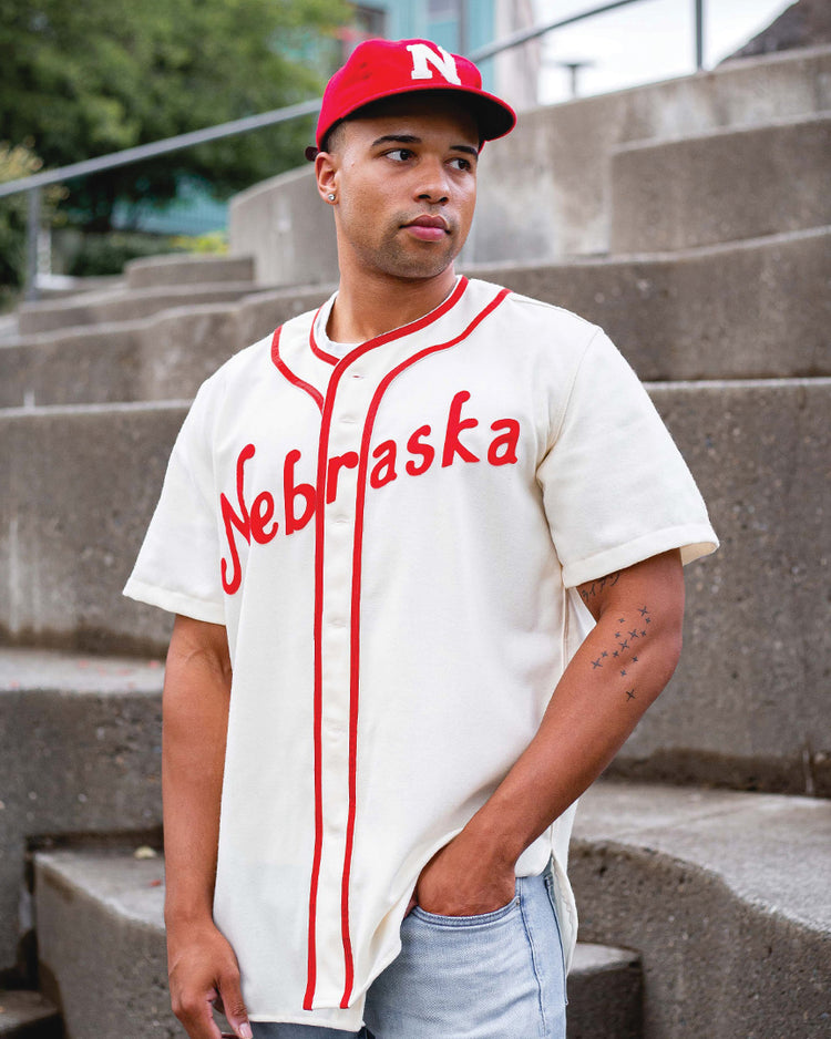 A man stands outdoors on concrete steps, looking to the side with one hand in his pocket, wearing an Ebbets Field Flannels University of Nebraska 1958 Vintage Ballcap and a white baseball jersey with Nebraska written on it.