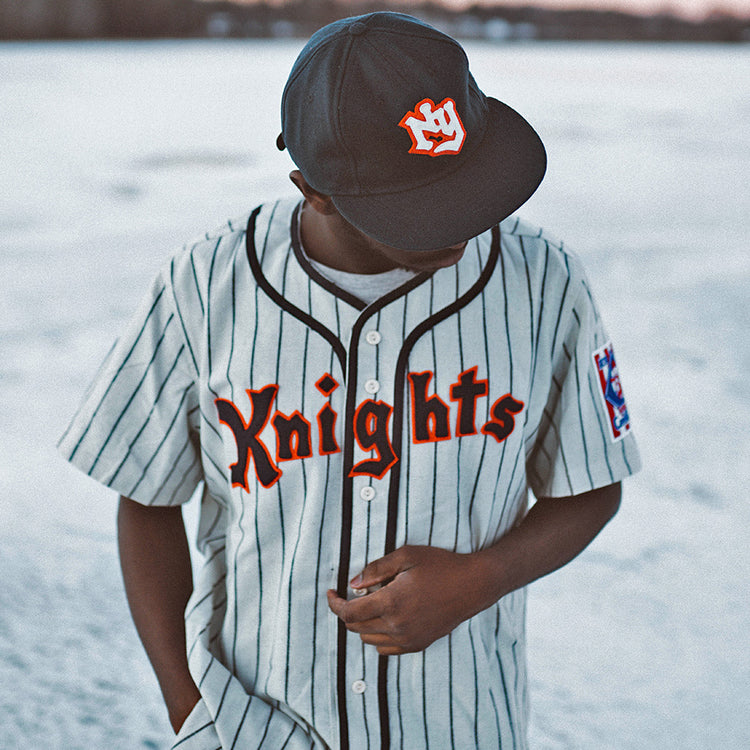 A person in an EFF New York Knights 1939 Home Jersey and black cap stands outdoors on snow or ice, looking down while adjusting their shirt.