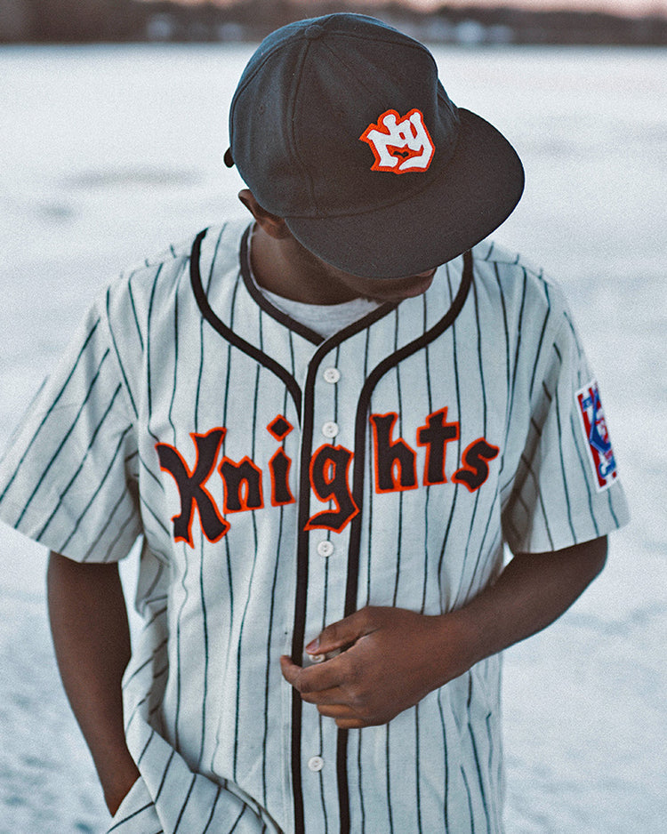 A person in an EFF New York Knights 1939 Home Jersey and black cap stands outdoors on snow or ice, looking down while adjusting their shirt.