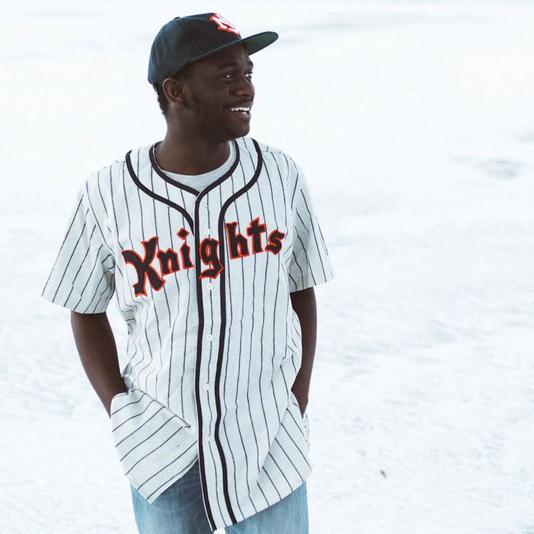 A person in an EFF New York Knights 1939 Home Jersey, featuring Knights across the front, pairs it with a black cap and jeans while standing outdoors, smiling and looking to the side with hands in their pockets.