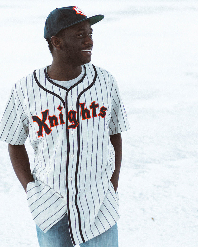 A person in an EFF New York Knights 1939 Home Jersey, featuring Knights across the front, pairs it with a black cap and jeans while standing outdoors, smiling and looking to the side with hands in their pockets.