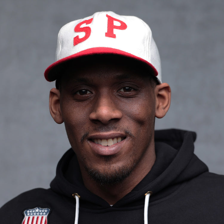 A man in a black hoodie and an EFF Nebraska State Prison 1923 Vintage Ballcap with red SP letters smiles at the camera against a plain gray background.