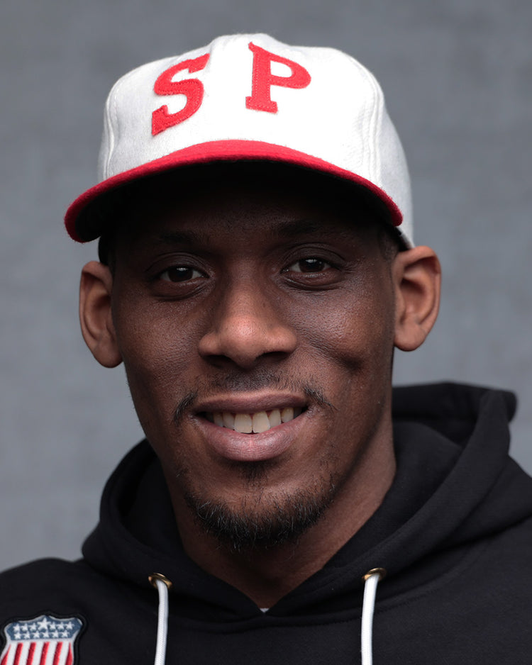 A man in a black hoodie and an EFF Nebraska State Prison 1923 Vintage Ballcap with red SP letters smiles at the camera against a plain gray background.