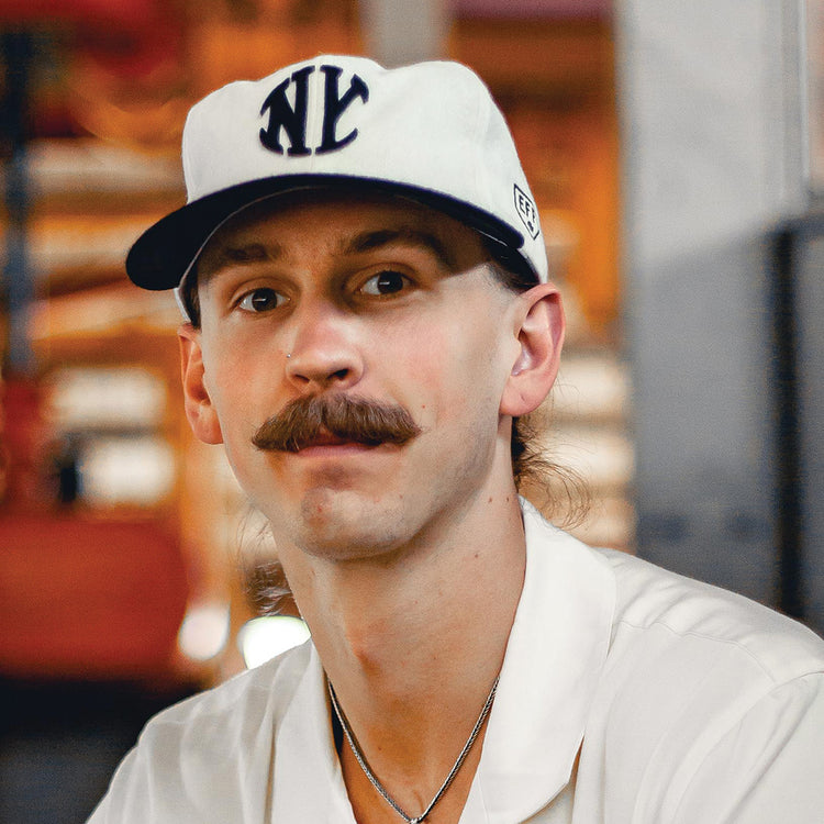 A man with a brown mustache wears an EFF New York Knickerbockers Vintage Inspired Ballcap—a white hat with bold NYC lettering—and a white shirt, gazing at the camera against a softly blurred, warm-toned background.