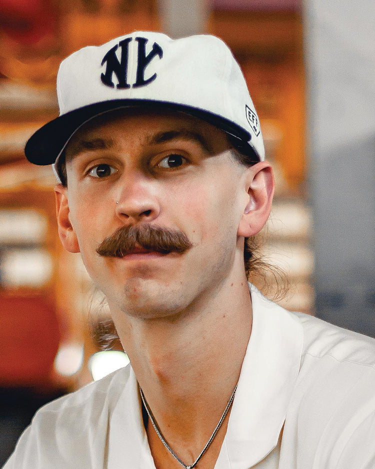 A man with a brown mustache wears an EFF New York Knickerbockers Vintage Inspired Ballcap—a white hat with bold NYC lettering—and a white shirt, gazing at the camera against a softly blurred, warm-toned background.