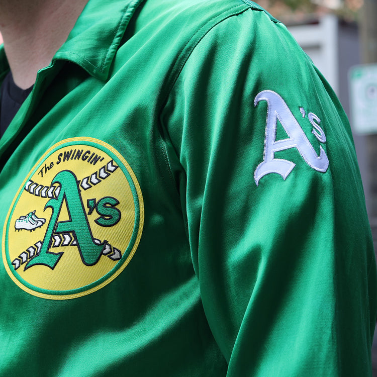 Someone wears the EFF Oakland As 1971 Satin Windbreaker, featuring a green satin finish, an Athletics As logo patch on the chest, and As lettering on the sleeve.