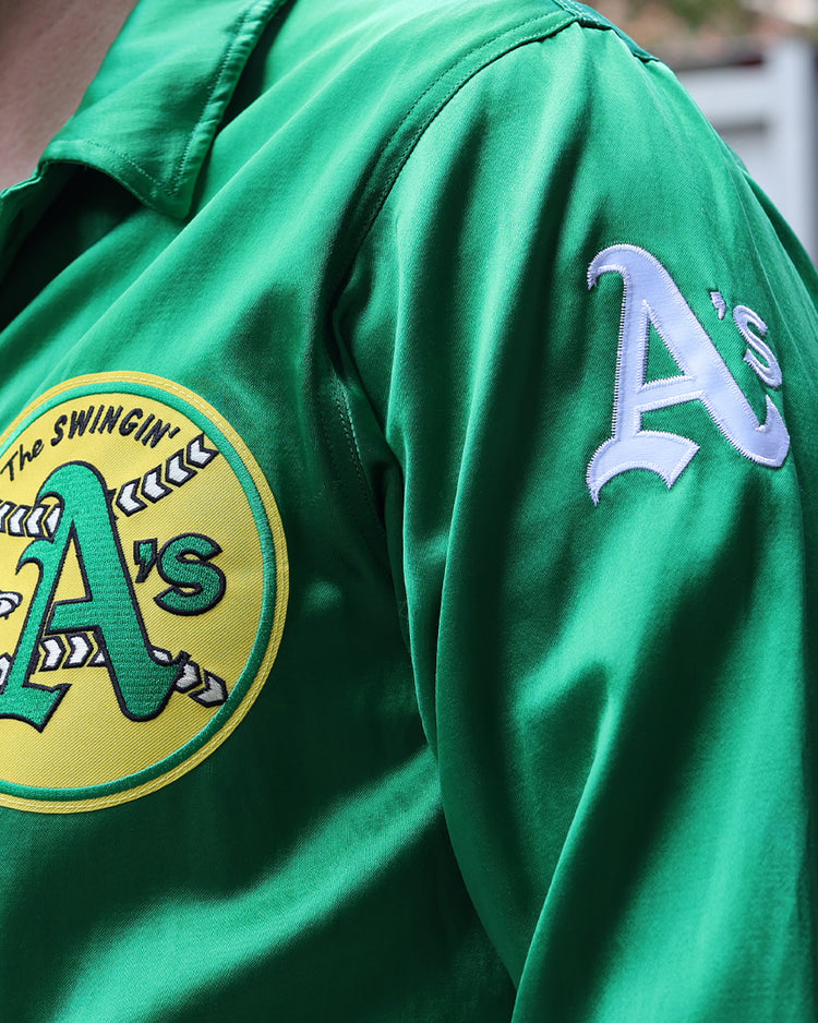 Someone wears the EFF Oakland As 1971 Satin Windbreaker, featuring a green satin finish, an Athletics As logo patch on the chest, and As lettering on the sleeve.