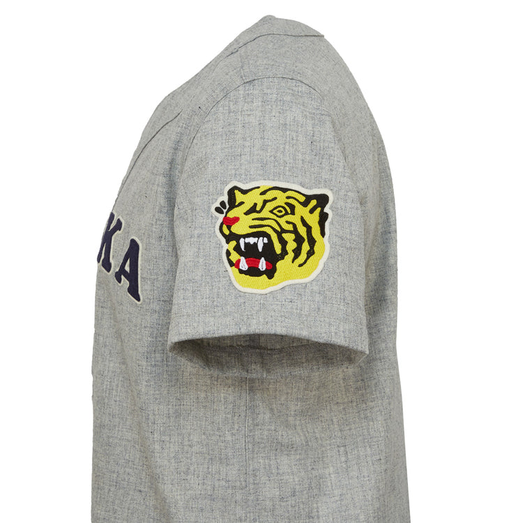 The EFF Osaka Tigers 1959 Road Jersey is a gray short-sleeve shirt inspired by Japanese baseball, featuring KA on the chest and a yellow tiger head patch on the sleeve, echoing classic Koshien Field jerseys.