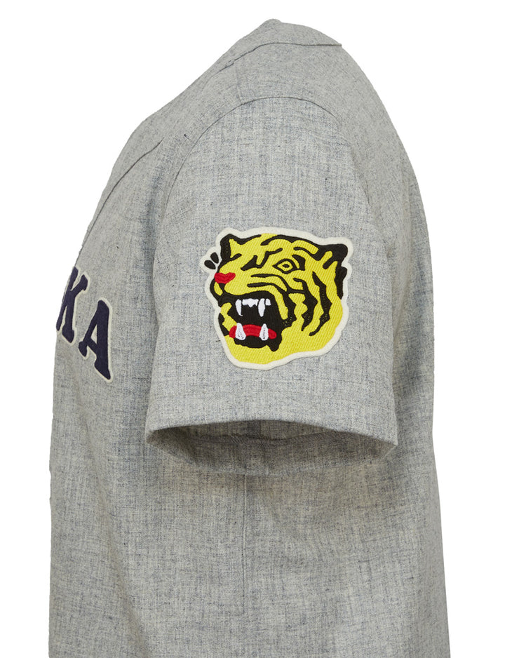 The EFF Osaka Tigers 1959 Road Jersey is a gray short-sleeve shirt inspired by Japanese baseball, featuring KA on the chest and a yellow tiger head patch on the sleeve, echoing classic Koshien Field jerseys.