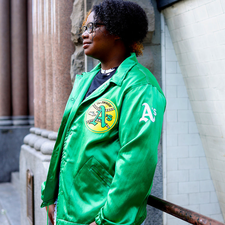A person with short curly hair and glasses wears the EFF Oakland As 1971 Satin Windbreaker, standing outdoors by a stone wall and tiled building, looking to the side.