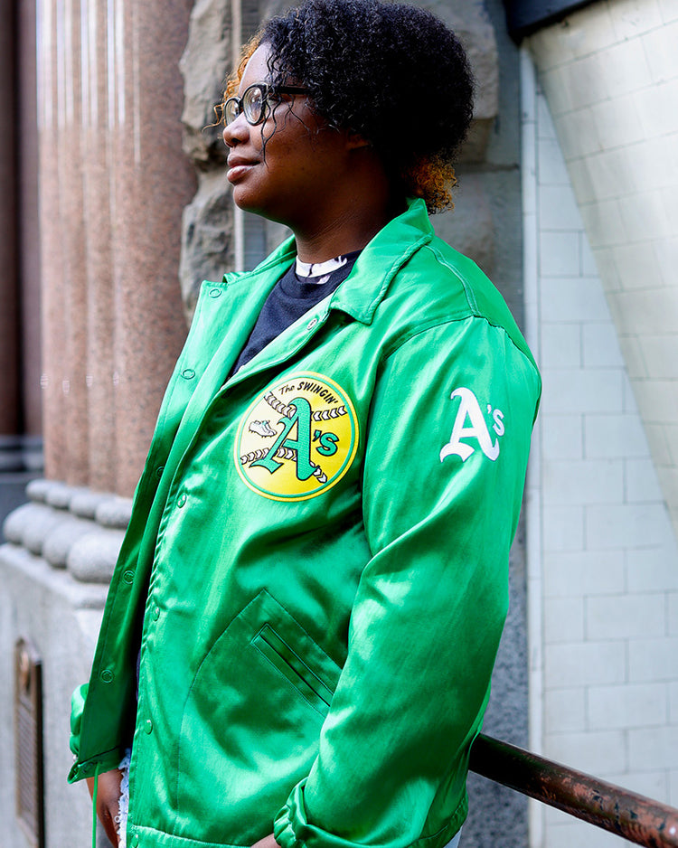 A person with short curly hair and glasses wears the EFF Oakland As 1971 Satin Windbreaker, standing outdoors by a stone wall and tiled building, looking to the side.