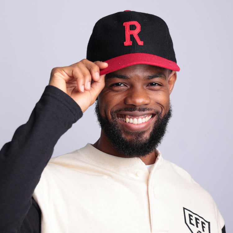 A bearded man smiles and tips his Rutgers University 1955 Vintage Ballcap by EFF, black and red with a large red R, while wearing a light shirt with black sleeves and an EFF patch. The background is plain light gray.