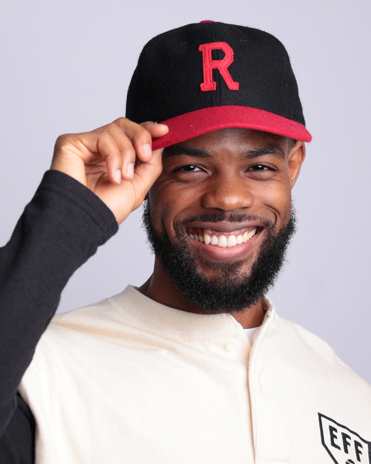 A bearded man smiles and tips his Rutgers University 1955 Vintage Ballcap by EFF, black and red with a large red R, while wearing a light shirt with black sleeves and an EFF patch. The background is plain light gray.