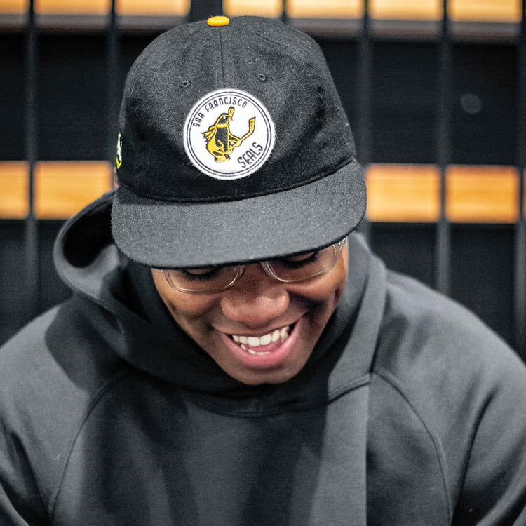 A person in a dark hoodie and glasses smiles while looking down, wearing the EFF San Francisco Seals 1963 Vintage Ballcap with a yellow and white logo. The background shows a black grid pattern.