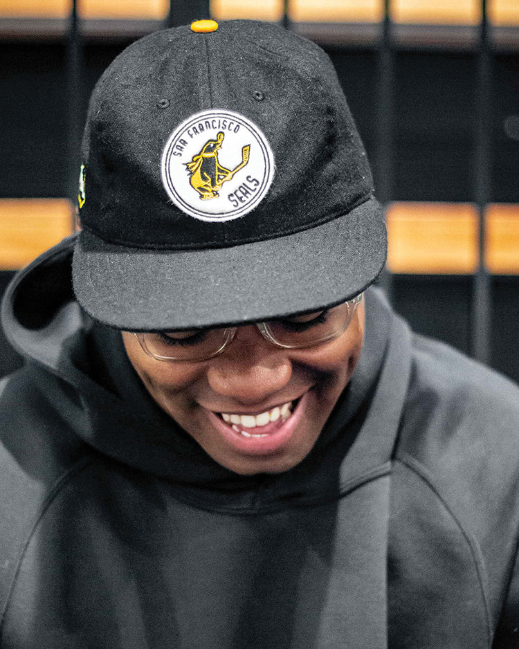 A person in a dark hoodie and glasses smiles while looking down, wearing the EFF San Francisco Seals 1963 Vintage Ballcap with a yellow and white logo. The background shows a black grid pattern.