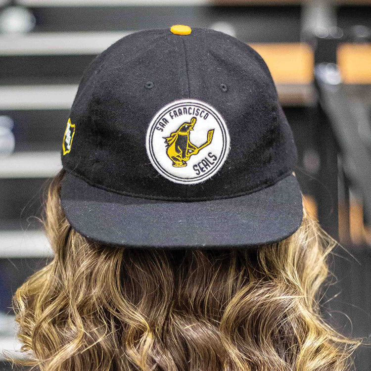 A person with wavy light brown hair wears the EFF San Francisco Seals 1963 Vintage Ballcap, featuring a circular patch with a yellow seal holding a bat. The background is blurred.
