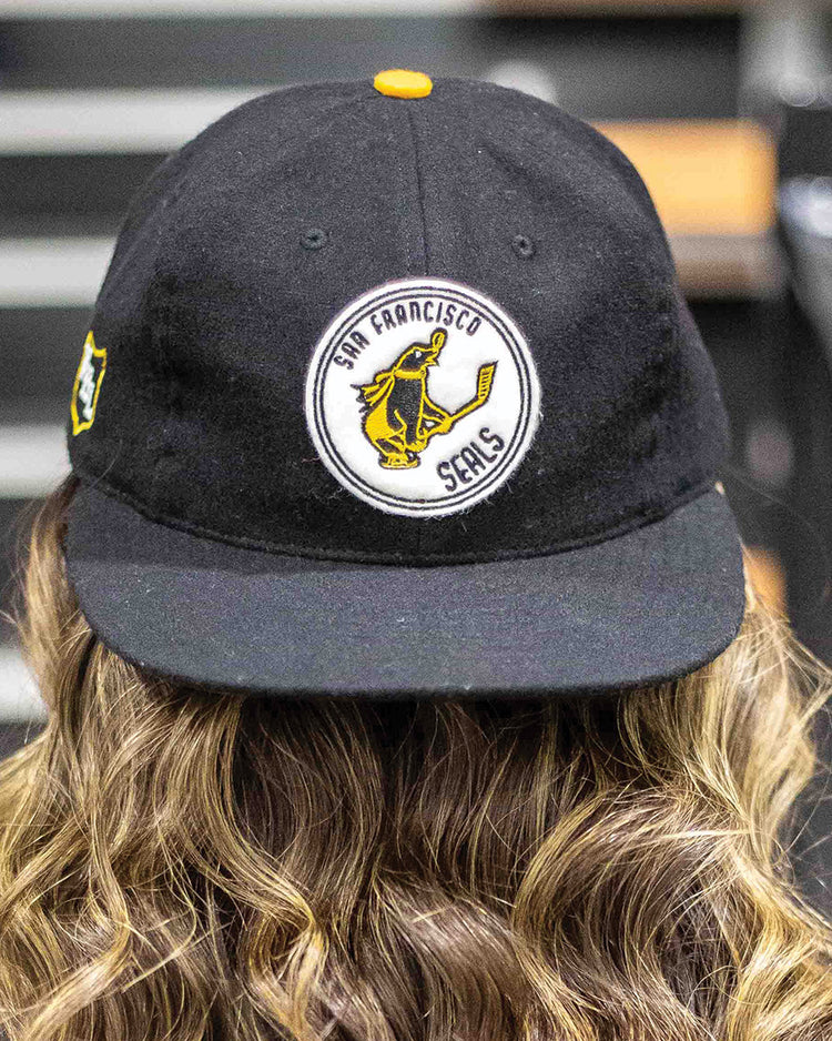 A person with wavy light brown hair wears the EFF San Francisco Seals 1963 Vintage Ballcap, featuring a circular patch with a yellow seal holding a bat. The background is blurred.