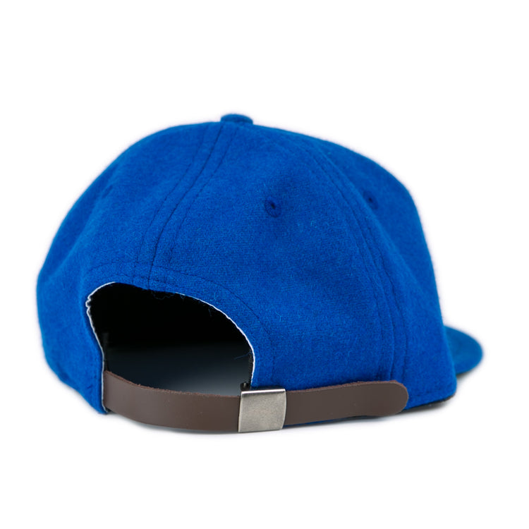 A close-up of the back of the blue EFF Southern Methodist University 1962 Vintage Ballcap shows its brown adjustable strap and metal buckle. The wool cap rests on a white surface.
