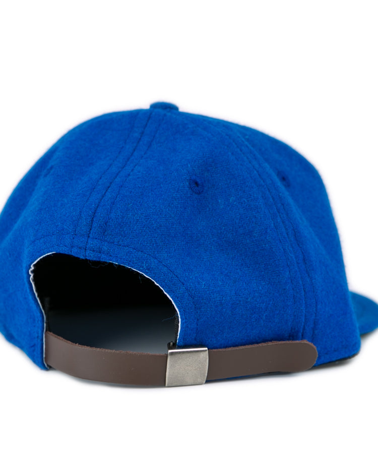 A close-up of the back of the blue EFF Southern Methodist University 1962 Vintage Ballcap shows its brown adjustable strap and metal buckle. The wool cap rests on a white surface.