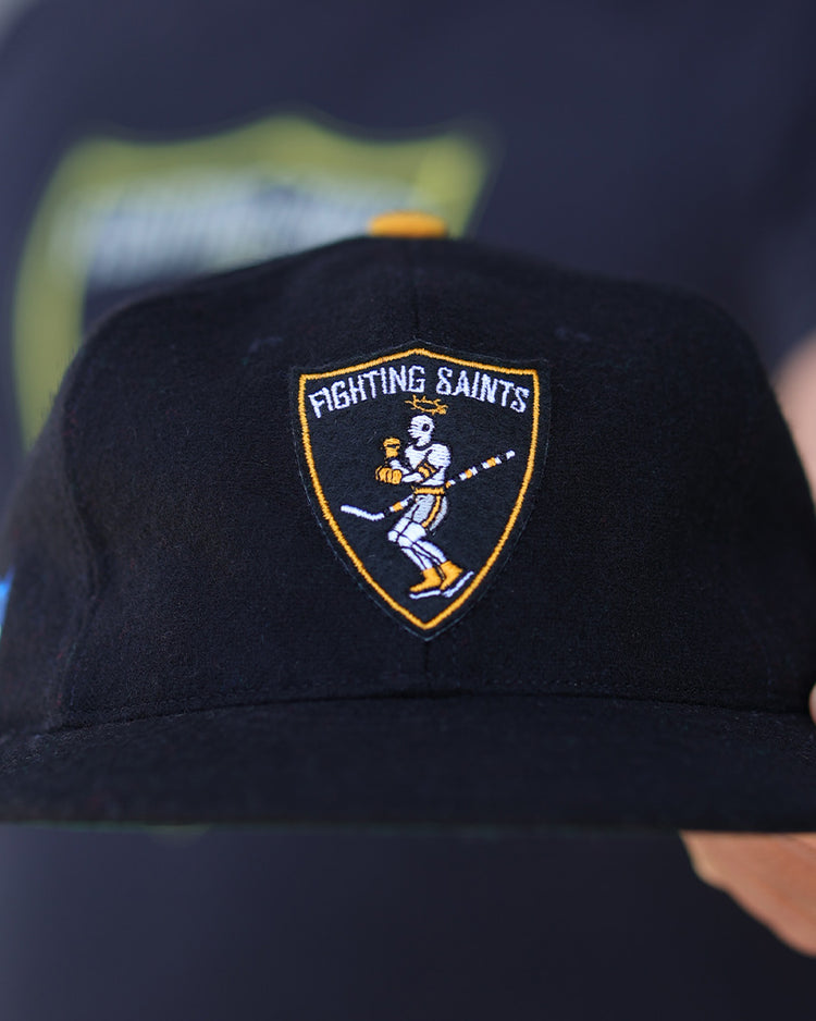 A person holds the EFF St. Paul Fighting Saints 1961 Vintage Ballcap, featuring a cartoon hockey player inside a yellow-bordered shield logo on the front, with a blurred background highlighting the cap.