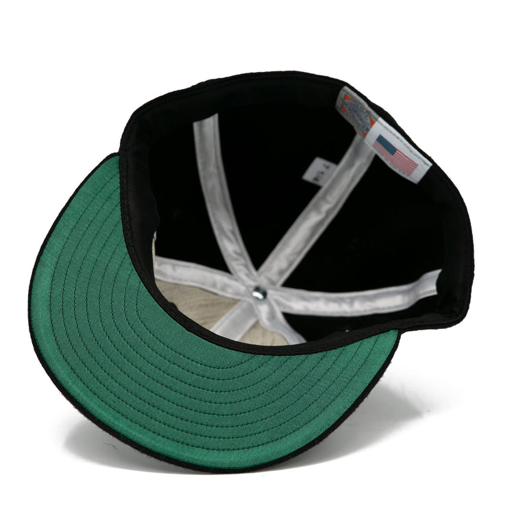 A look inside the EFF Brandon Wheat Kings 1955 Vintage Ballcap reveals black wool, a green brim underside, white sweatband, visible seams, and an inner American flag tag—reflecting classic hockey style.