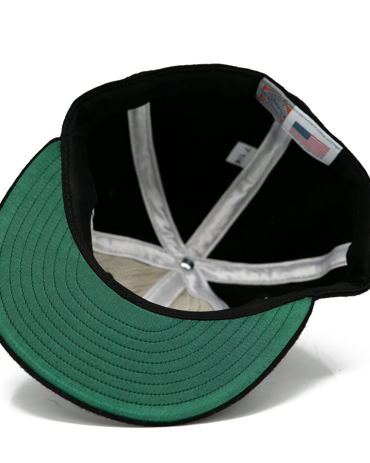 A look inside the EFF Brandon Wheat Kings 1955 Vintage Ballcap reveals black wool, a green brim underside, white sweatband, visible seams, and an inner American flag tag—reflecting classic hockey style.