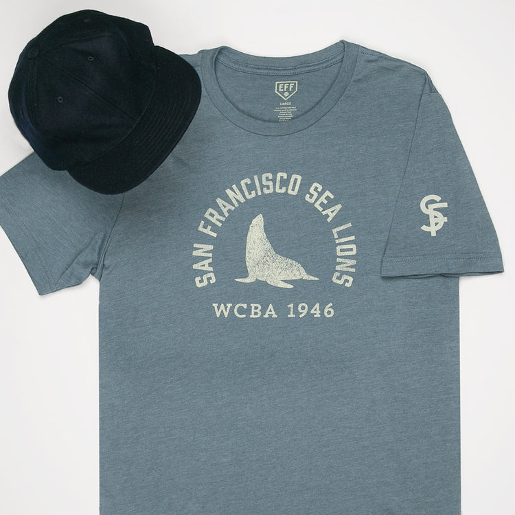 The EFF San Francisco Sea Lions 1946 T-Shirt features premium blue cotton with a sea lion graphic and WCBA 1946 text, inspired by the West Coast Baseball Association, shown paired with a black baseball cap.