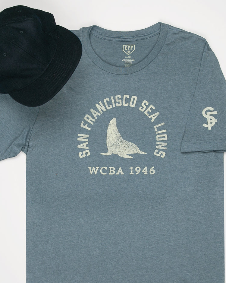 The EFF San Francisco Sea Lions 1946 T-Shirt features premium blue cotton with a sea lion graphic and WCBA 1946 text, inspired by the West Coast Baseball Association, shown paired with a black baseball cap.