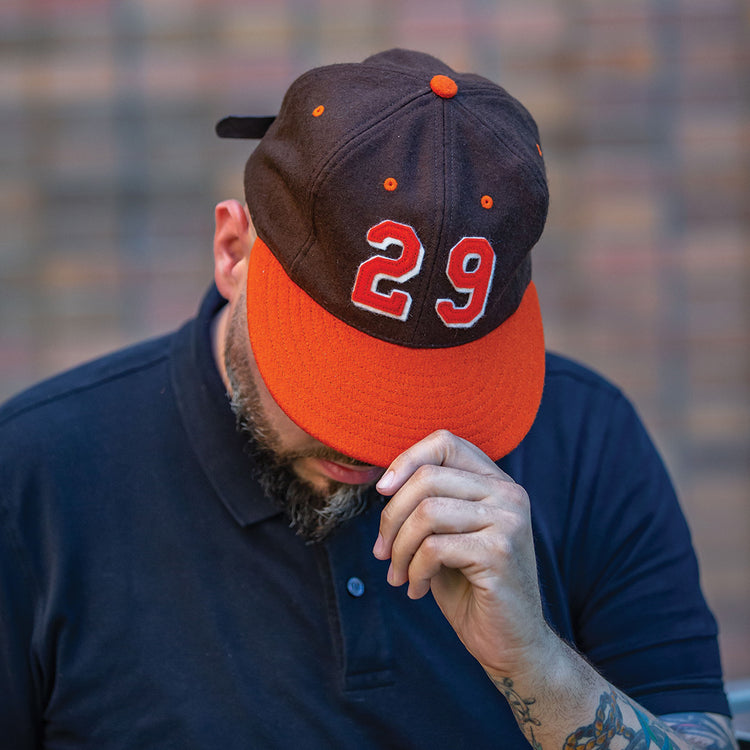 A person in a navy blue shirt holds the brim of an Ebbets Field Flannels Satchel Paige 1952 Signature Series Ballcap—orange and black with “29” on the front. Their head is down, partially obscuring their face against a blurred brick wall.