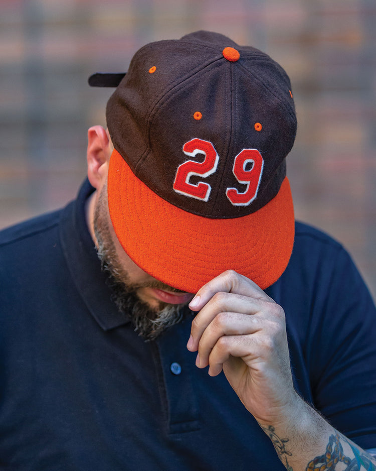 A person in a navy blue shirt holds the brim of an Ebbets Field Flannels Satchel Paige 1952 Signature Series Ballcap—orange and black with “29” on the front. Their head is down, partially obscuring their face against a blurred brick wall.