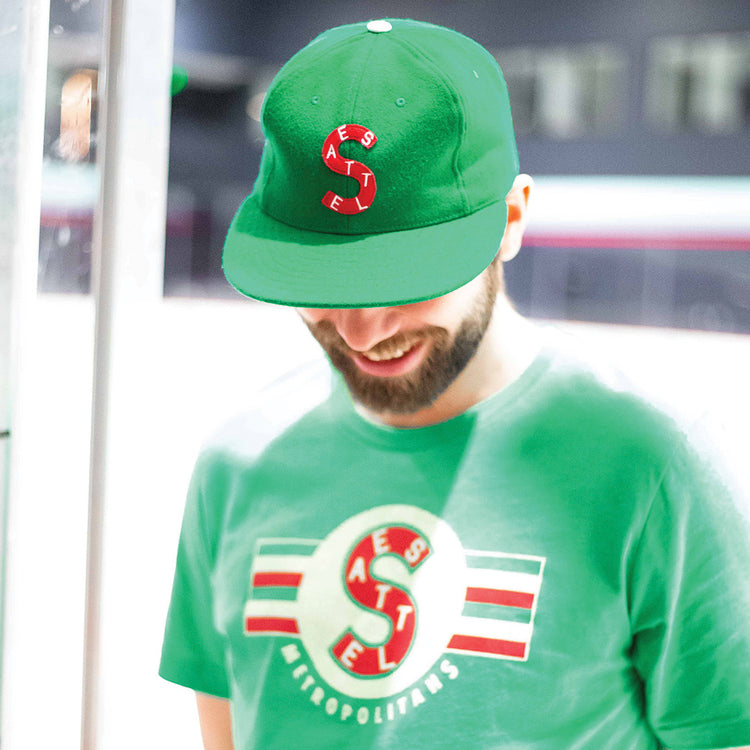 A man wearing an EFF Seattle Metropolitans 1917 Vintage Ballcap—a green cap with a red and white “S” logo—smiles while looking down. He also wears a matching green T-shirt; the background is blurred.