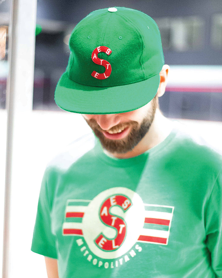 A man wearing an EFF Seattle Metropolitans 1917 Vintage Ballcap—a green cap with a red and white “S” logo—smiles while looking down. He also wears a matching green T-shirt; the background is blurred.